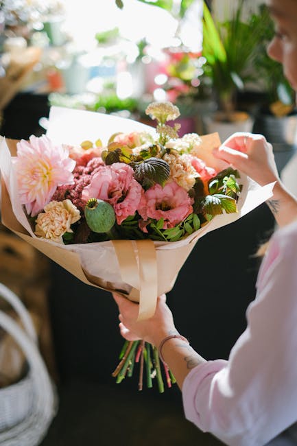 A person holding a large, vibrant floral bouquet featuring pink peonies, cream-colored carnations, and small white and purple filler flowers, all wrapped in light beige paper with a satin ribbon. The 