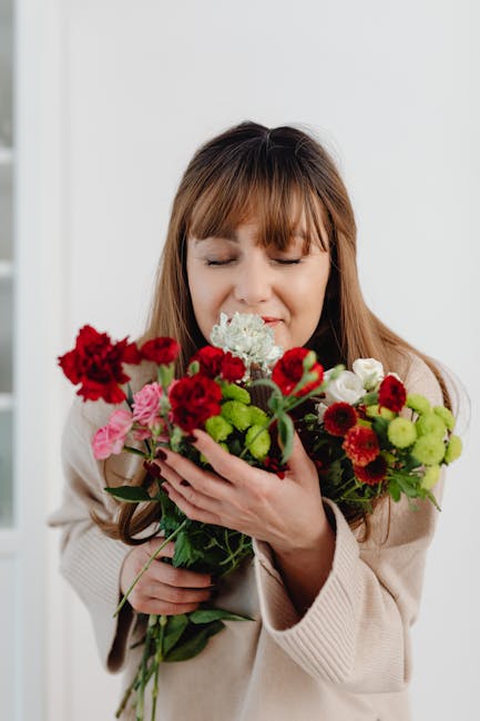 A woman with light brown hair and bangs, dressed in a beige sweater, is holding a vibrant bouquet of fresh flowers in both hands. The bouquet features a variety of blooms, including deep red and pink 
