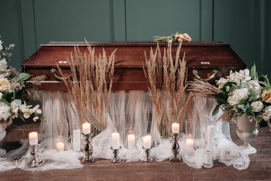 A funeral arrangement featuring a polished wooden casket surrounded by an elegant display of fresh flowers including white roses, lilies, and soft pastel peonies, accented with green foliage. Dried pa