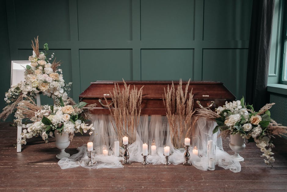 A floral arrangement dedicated to funeral or memorial services, featuring two large white vases filled with white roses, peonies, and eucalyptus, with accents of beige pampas grass. These vases are po