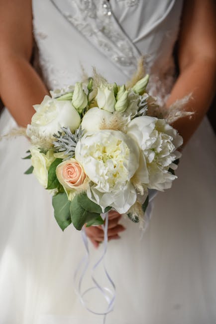 A bride holding a bouquet arrangement featuring predominantly white flowers, including large peonies, roses, and lisianthus, complemented by grayish-green foliage and delicate, wispy accents. The bouq