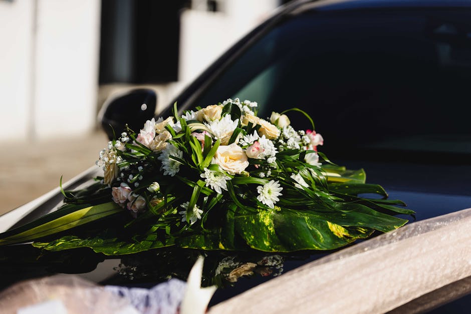 A delicate floral arrangement featuring white roses, daisies, and small pink blossoms, accented with lush green foliage, is placed on the hood of a black car, likely prepared for a wedding or special 