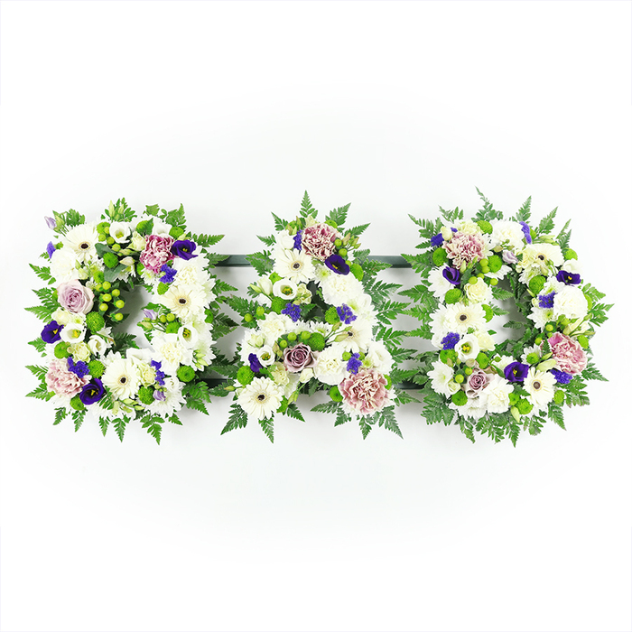 This image shows a classic funeral flower tribute spelling out DAD in three bold, floral letters, perfectly aligned in a row and photographed against a simple white backdrop that lets every bloom speak for itself. Each letter is expertly packed with pristine white flowers: domed chrysanthemums, daisy-like white gerberas with soft centres, and elegant white lisianthus and spray roses, layered together so that the surface appears plush and continuous. The flowers are placed so closely that the letters look almost sculpted from petals, with the rounded edges and open spaces of the D and A carefully defined. Around these white blooms, rich green foliage - notably delicate fern fronds and rounded leaves - creates a verdant border that frames the word and adds depth. Tucked within the greenery are pops of bright green button chrysanthemums or berries, giving a fresh, youthful note that balances the formality of the white. Across the letters, selective touches of colour are woven: deep violet and purple flowers reminiscent of pansies or small chrysanthemums, as well as softly toned lavender roses and pale mauve carnations. These accents lend a sense of quiet luxury and gentle warmth rather than overpowering the design. The composition feels calm and considered, the kind of tribute families in Maidenhead might choose for a funeral service at a local church or crematorium, or to rest in a cemetery near familiar streets and riverside walks. It is a personalised floral message that honours a father's memory with grace, representing love, respect, and the enduring bond between parent and child.