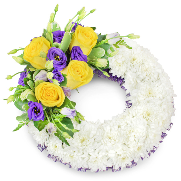 A refined, circular sympathy wreath photographed on a simple white field-its clean presentation emphasizing the handcrafted detail and the respectful mood of the piece. The majority of the wreath's circumference is built from tightly clustered white spray chrysanthemums, whose small, layered petals create a plush, even surface resembling a soft, clouded band. Against that pure white foundation, an eye-catching floral spray sits on the upper left: several large yellow roses bloom fully, their petals unfurling in warm, sunlit tones that feel comforting and familiar. Nestled alongside these roses are deep purple lisianthus, their cup-shaped, ruffled blossoms lending a sense of elegance and calm. A sprinkling of pale lavender lisianthus buds and tender green shoots provides depth and the suggestion of quiet renewal. Mixed foliage-shiny rounded leaves, delicate fern fronds, and a few variegated leaflets-frames the colour, offering contrast and graceful movement that prevents the composition from feeling static. Along both the outer and inner edges of the chrysanthemum ring a slender hint of dark purple mesh or crinoline is visible, quietly echoing the lisianthus hues and providing a polished edge. The open centre of the wreath maintains its traditional loop, a symbolic circle of remembrance. As a florist who frequently prepares tributes for services near Maidenhead Railway Bridge and local chapels, I find this design speaks gently of peace and enduring affection, its scent, texture, and palette thoughtfully chosen to convey condolence.
