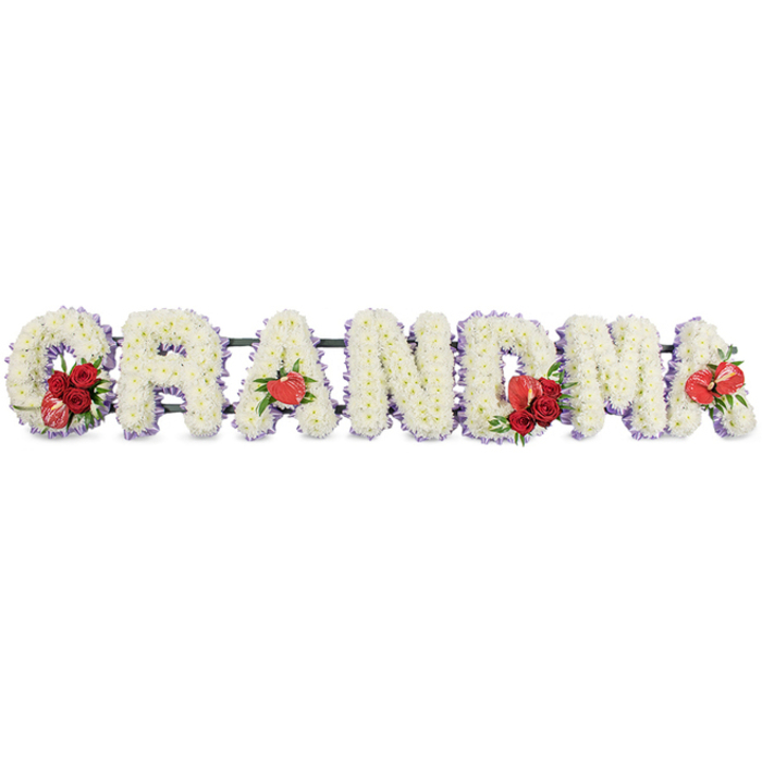 A beautifully composed sympathy tribute in this photograph spells out GRANDMA in bold, upright letters, set horizontally and photographed against a clear white backdrop so the craftsmanship is easy to imagine. Each character is entirely filled with clusters of white chrysanthemums, tightly arranged so that no gaps show, giving the letters a plush, cushioned appearance reminiscent of soft clouds or freshly fallen petals on the paths of Kidwells Park in Maidenhead. The flowers' small, rounded heads catch the light and create gentle shadows that emphasise the contours of each letter. Around the outside edges, a trim of pale lavender or lilac ribbon frames the white blooms, lending a delicate contrast and a feeling of calm spirituality without overwhelming the simplicity of the design. Touches of vivid red are incorporated as focal points of love and remembrance: the G is enriched by a spray of deep red roses combined with an eye-catching variegated red-and-white calla lily nestled into its lower curve. The first A is highlighted with a solitary heart-shaped red anthurium at its base, its waxy surface and elongated spadix adding an interesting textural contrast. At the centre, the M carries another cluster of red roses accompanied by a second red anthurium, while the final A features a last, single red anthurium, visually linking the beginning and end of the word. Beneath the blossoms, a dark green framework supports each letter, slightly visible yet discreet. This carefully balanced arrangement conveys both serenity and devotion, the kind of personalised funeral flowers a local Maidenhead family might order to lie beside a casket or at a memorial service in honour of a much-loved grandmother.