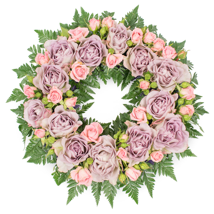 A full, round sympathy wreath is captured from directly above, resting on a pure white surface that highlights its colours and careful construction. The arrangement forms a smooth, unbroken circle, densely filled with large roses in a muted palette of dusty lavender, antique mauve, and soft grey-purple. Their petals are layered and abundant, curling inwards in delicate, rose-like swirls that give the wreath a sense of quiet luxury. Tucked among these statement blooms are numerous small spray roses in a gentle pastel pink, some just opening, some still in bud, bringing lightness and a tender, youthful energy to the design. The flowers emerge from a substantial backing of dark green fern fronds, whose finely divided, arching leaves spill slightly beyond the circular outline to create a softly feathered edge. Deeper filler foliage and small, shadowy buds are interlaced between the roses, providing contrast and visual depth, much like the shaded paths under the trees in Maidenhead's Grenfell Park. The overall effect is harmonious and serene, with cool purples and warm blush tones balanced against the lush greenery. This wreath feels suited to a funeral, memorial service, or graveside farewell anywhere in the Maidenhead area, whether delivered to a family home along the Thames or to a local chapel. The composition suggests experienced hands guiding every stem into place, mindful of symmetry yet allowing for natural variation. It reads as a circle of remembrance and respect, a floral gesture meant to comfort mourners while quietly celebrating a cherished life.