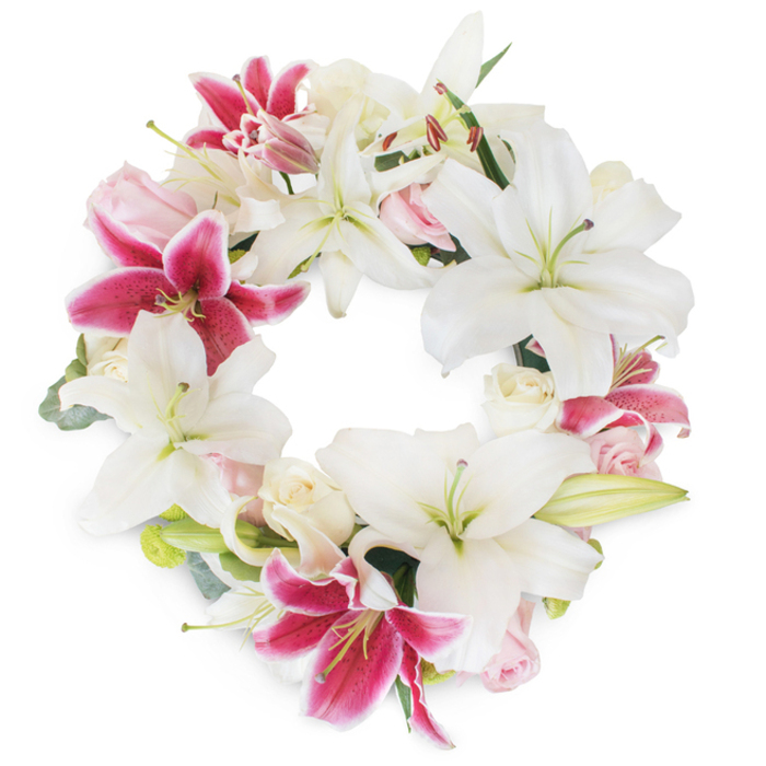 Taking pride of place on a pure white background, this sumptuous memorial wreath is a testament to Maidenhead's tradition of gentle remembrance. Large, star-studded white lilies dominate the composition, each petal unfurling gracefully, their pristine hue evoking both peace and new beginnings. Striking fuchsia-pink lilies, with captivating magenta tones and speckled centers, provide a vivid contrast, reflecting the vibrant character found in Maidenhead's riversides and historic High Street. Nestling between these are clusters of delicate blush and creamy roses-some tightly closed, others blossoming into soft fullness-offering a subtle texture and a touch of romance. Muted green foliage, both glossy and silvered, adds natural structure and depth, peeking from behind the main blooms and recalling the tranquil green spaces cherished throughout the area. Every detail, from the shimmering pollen on the lilies' stamens to the velvety layers of each rose petal, is arranged with intention by Maidenhead's experienced florists. The circle is complete and uninterrupted, signifying everlasting love and remembrance-a fitting centrepiece for a funeral or a celebration of life. Harmonious in colour and sensitive in design, this floral wreath is an eloquent way to extend sympathy, honor a loved one's journey, or express support to families around Maidenhead's community, where heartfelt tributes find their home.