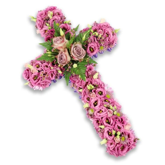 Set against a crisp white background with a subtle, soft-edged shadow, a beautifully composed floral cross fills the frame, its entire surface densely covered in fresh blossoms in shades of pink, mauve, and dusky lavender. The craftsmanship suggests an experienced Maidenhead florist has taken great care, placing each flower close to its neighbour so that the cross feels like a single, flowing carpet of petals. At the intersection of the horizontal and vertical arms, three large roses serve as a gentle focal point, their petals in a muted dusty rose and light mauve hue unfurling in layered spirals that catch the light. These focal roses rest in a nest of feathery green fern fronds and smaller bright green leaves, which provide a vivid contrast and evoke the soft greenery seen around Maidenhead's riverside paths and local churchyards. Extending outward from the centre, the arms and base of the cross are lavishly filled with lisianthus blooms in soft pink, mid-pink, and lilac tones, many with darker, velvety centres, adding an almost watercolor-like depth. Threaded between these larger blooms are tiny clusters of pink and purple filler flowers, delicate white blossoms tipped with pale yellow centres, and occasional buds of light green, hinting at fresh growth amidst remembrance. Along some edges, slim lines of darker purple ribbon or structural fabric briefly appear, framing the design and emphasizing the cross shape without overpowering the blossoms. The mood of the arrangement is one of reverent peace and heartfelt condolence, an ideal floral cross for a funeral or memorial service in Maidenhead, whether placed beside the coffin, at the front of the church, or at a graveside to honour a cherished life.