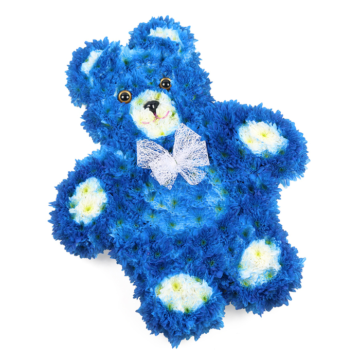 In this detailed close-up, a floral teddy bear tribute lies slightly on its side against a flawless white background, its colours and textures taking centre stage. The entire silhouette of the bear-from its rounded ears to its little feet-is created from a dense carpet of royal blue blossoms, probably small chrysanthemums or similar multi-petalled flowers, each bloom nestled so tightly that the surface looks like soft, luxurious fur. Delicate clusters of cream and pale yellow flowers mark the inner ears, circular snout, and four oval paw pads, giving the bear clearly defined features and a tender, toy-like charm. The eyes are made from shiny, dark brown or amber elements that catch the light, while a small black triangular nose and a gently curved stitched-style mouth add a subtle, comforting smile. Around the neck, a sheer white bow made from fine mesh or organza sits lightly on the blooms, its translucent loops and trailing ends adding an elegant, almost angelic touch. The lighting is bright and even, with no shadows to distract from the intricate floral work, so the viewer can almost feel the soft, cool petals beneath their fingertips. This arrangement, created in the style of a flat-backed tribute, feels ideal for resting on a coffin or display stand at a funeral service in Maidenhead, perhaps in a chapel near the River Thames or a crematorium serving local families. The deep blue colour suggests calm, remembrance, and everlasting love, making this floral teddy bear a poignant way to honour the memory of a child or to send a message of gentle comfort when words are hard to find.